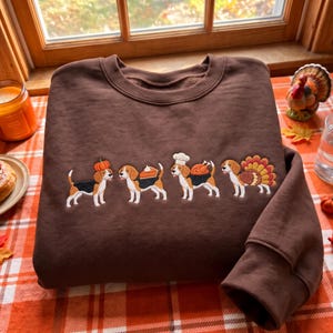 May include: A dark brown crewneck sweatshirt featuring an embroidered design of four beagle dogs. The first dog wears a pumpkin, the second carries a slice of pie, the third wears a chef hat with a roasted turkey, and the fourth has turkey feathers.