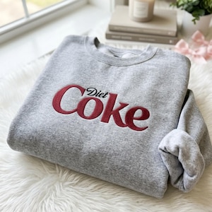 Op de afbeelding: Een grijs gemêleerd sweatshirt met ronde hals en de geborduurde tekst Diet Coke in een donkerrode kleur op de borst. Het kledingstuk heeft lange mouwen met geribbelde boorden en een geribbelde halslijn in een klassiek pullover-ontwerp.