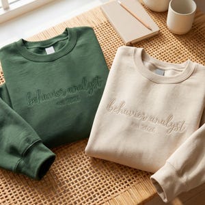 Op de afbeelding: Een donkergroene sweater en een beige sweater, beide met een ronde hals en lange mouwen. Elk kledingstuk is voorzien van de geborduurde tekst "behavior analyst" in cursief schrift, met daaronder de kleinere gedrukte tekst "est 2026".
