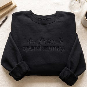 May include: A black long-sleeve sweatshirt featuring the text "I do pilates & spend money." embroidered in a matching black thread across the chest. The garment has a crew neckline and long sleeves with ribbed cuffs, showcasing a monochromatic design.
