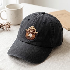 Könnte beinhalten: Eine anthrazitfarbene Baseballkappe mit einem gestickten Aufnäher eines braunen Bären, der einen beigen Rangerhut trägt. Das Wort RESIST ist in dunkelbraunen Buchstaben auf der Vorderseite der Krempe des Rangerhuts des Bären gestickt.