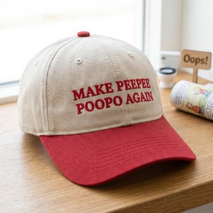 May include: A beige baseball cap with a red brim and a red button on top. The front of the cap features the embroidered red text "MAKE PEEPEE POOPO AGAIN" in a bold, sans-serif font. The cap has stitched eyelets on the crown panels.