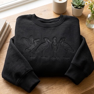 Peut inclure: Un sweat-shirt noir à manches longues avec un motif brodé ton sur ton représentant quatre cigognes à bec en sabot dans diverses poses sur la poitrine. Le vêtement présente un col rond, des poignets et un ourlet côtelés, avec des détails précis.