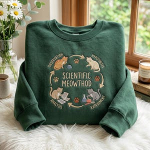 Op de afbeelding: Een donkergroene sweater met ronde hals en een cirkelvormige afbeelding van vier katten die wetenschappelijke taken uitvoeren. In het midden staat de tekst "SCIENTIFIC MEOWTHOD", omringd door de woorden "OBSERVATION", "HYPOTHESIS", "EXPERIMENT" en "ANALYSIS".