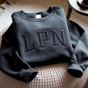 Può includere: Una felpa grigio scuro con le lettere "LPN" in un design a blocchi in rilievo. Sotto le lettere, le parole "LICENSED PRACTICAL NURSE" sono stampate. La felpa è piegata su una superficie beige intrecciata.