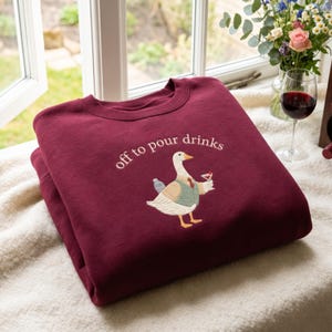 May include: A maroon crewneck sweatshirt featuring an embroidered graphic of a white goose wearing a tan waistcoat and red tie. The goose holds a cocktail glass and carries a shaker. The text "off to pour drinks" is embroidered above the goose.