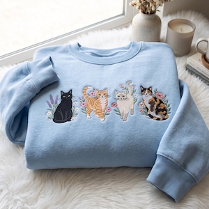 Könnte beinhalten: Ein hellblaues Sweatshirt mit Rundhalsausschnitt und einer Stickerei von vier Katzen in einer Reihe. Das Motiv zeigt eine schwarze Katze, eine orangefarbene getigerte Katze, eine cremefarbene Katze und eine Glückskatze, umgeben von Blumen.