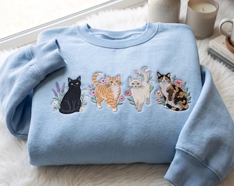 Op de afbeelding: Een lichtblauw sweatshirt met ronde hals en een geborduurd ontwerp van vier katten op een rij. De katten omvatten een zwarte kat, een oranje cyperse kat, een lichtcrème kat en een lapjeskat, omringd door kleine kleurrijke bloemen en bladeren.