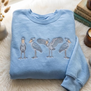 May include: A light blue crewneck sweatshirt featuring an embroidered design of four shoebill storks in various poses. The birds are depicted in shades of grey and blue with tan beaks, showcasing different stances including one with wings spread wide.