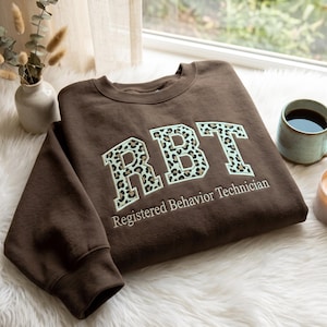 May include: A dark brown crewneck sweatshirt featuring the letters RBT in a large, block-style font with a light green and black leopard print pattern. Below the letters, the text Registered Behavior Technician is embroidered in white.
