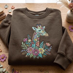 May include: A dark brown crewneck sweatshirt featuring a central embroidered design of a giraffe head and neck surrounded by a colourful arrangement of various flowers and leaves in shades of pink, yellow, blue, and green.