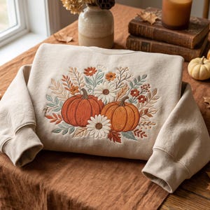 Op de afbeelding: Een beige sweater met ronde hals en een borduursel op de voorzijde. Het ontwerp toont twee oranje pompoenen omringd door witte en oranje bloemen, groene bladeren en dunne takjes, wat zorgt voor een gedetailleerd botanisch arrangement.