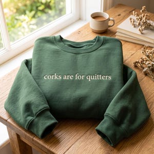 Puede incluir: Una sudadera de cuello redondo en color verde bosque con el texto bordado "corks are for quitters" en color crema claro en el pecho. La prenda tiene mangas largas con puños acanalados y un cuello acanalado, confeccionada en tela suave.