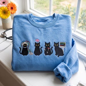 Könnte beinhalten: Ein hellblaues Sweatshirt mit einer Stickerei von vier schwarzen Katzen. Die erste Katze trägt eine EEG-Haube mit bunten Kabeln, die zweite hat ein Gehirn-Symbol darüber, die dritte hält einen Reflexhammer und die vierte sitzt neben einem Monitor.