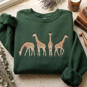 May include: A dark forest green crewneck sweatshirt featuring an embroidered design of four giraffes in various poses. The giraffes are depicted with brown and tan spotted patterns, showcasing one grazing, two standing, and one facing forward.