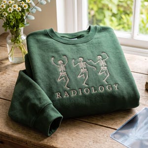Peut inclure: Un sweat-shirt à col rond vert forêt avec une broderie représentant trois squelettes dansants en fil blanc. Sous les squelettes, le mot RADIOLOGY est brodé en lettres capitales blanches. Le vêtement possède des manches longues et des poignets côtelés.