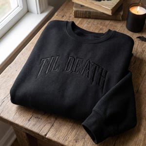 May include: A black crewneck sweatshirt featuring the text TIL DEATH embroidered in a matching black thread across the chest in a gothic-style font. The garment has long sleeves with ribbed cuffs and a ribbed neckline, showcasing a monochromatic design.
