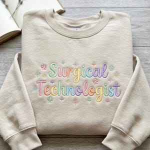 Könnte beinhalten: Ein beigefarbenes Sweatshirt mit Rundhalsausschnitt und dem gestickten Text "Surgical Technologist" in einer mehrfarbigen Pastellschrift. Das Design ist mit kleinen gestickten Blumen und Sternformen in Rosa, Gelb, Blau und Grün verziert.