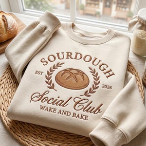 Può includere: Felpa color crema con ricamo marrone. Presenta le parole "SOURDOUGH Social Club", una pagnotta e una corona d'alloro. Il testo "WAKE AND BAKE" è sotto. La felpa è su un tappetino intrecciato.