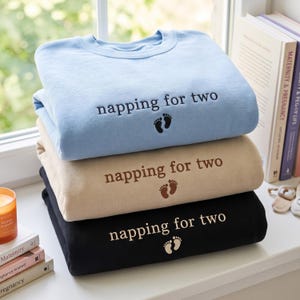 May include: A stack of three crewneck sweatshirts in light blue, beige, and black. Each sweatshirt features the embroidered text 'napping for two' in a lowercase font, accompanied by a small graphic of two baby footprints centred below the text.