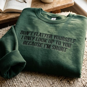 Pode incluir: Um moletom verde escuro com o texto bordado: "DON'T FLATTER YOURSELF I ONLY LOOK UP TO YOU BECAUSE I'M SHORT". O moletom está dobrado sobre uma superfície texturizada, com um livro e óculos ao fundo.