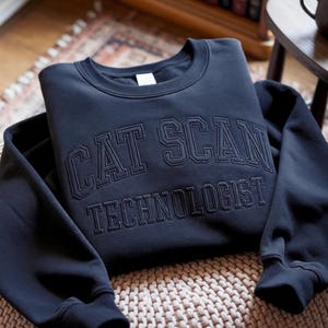 Op de afbeelding: Een donkerblauw sweatshirt met ronde hals en de tekst CAT SCAN TECHNOLOGIST geborduurd in bijpassend donkerblauw garen op de borst in een vetgedrukt lettertype in universiteitsstijl, met een geribbelde kraag en boorden.