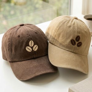 Puede incluir: Dos gorras de béisbol, una de color marrón oscuro y otra de color tostado claro, cada una con un diseño bordado de tres granos de café en el panel frontal. Las gorras tienen una corona estructurada, visera curva y ojales cosidos.