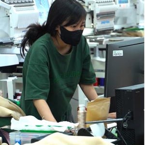 May include: A dark green short-sleeved t-shirt with dark green lettering on the chest, worn with a black face mask. The garment is in a workspace with industrial embroidery machines, a computer monitor, and various sewing supplies.