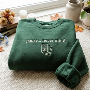 Könnte beinhalten: Ein waldgrüner Rundhals-Pullover mit dem gestickten Text "pause... never mind." in hellbeigem Garn. Unter dem Text befindet sich eine Stickerei von zwei Mahjong-Steinen, wobei der obere Stein das chinesische Schriftzeichen für sechs zeigt.
