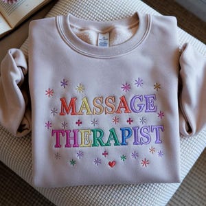 Op de afbeelding: Een lichtbeige sweatshirt met de woorden "MASSAGE THERAPIST" geborduurd in regenboogkleuren. Het ontwerp bevat sneeuwvlokken en een klein rood hart. De trui is netjes op een patroon oppervlak gevouwen.