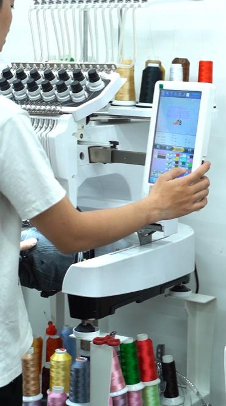 May include: A white industrial embroidery machine featuring a digital touchscreen interface, multiple thread spools in various colors, and a needle bar assembly. The machine is set up for fabric embroidery with a clear view of the control panel.