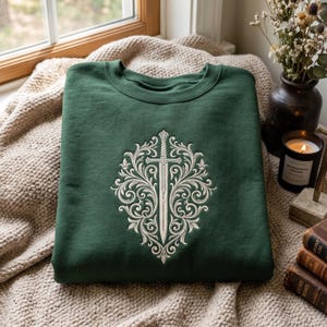 Pode incluir: Uma camisola de gola redonda em verde floresta com um bordado central em tom creme, apresentando uma espada vertical rodeada por padrões florais simétricos e intrincados. O design está posicionado no centro do peito.