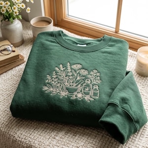 May include: A dark forest green crewneck sweatshirt featuring a central cream-coloured embroidered design. The embroidery depicts a botanical arrangement of various herbs, flowers, a mortar and pestle, and two small glass apothecary bottles.
