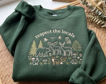 May include: A dark green crewneck sweatshirt featuring an embroidered design with the text "respect the locals" above a forest scene. The embroidery depicts a bear, moose, deer, owl, fox, squirrel, rabbit, hedgehog, trees, and mushrooms.