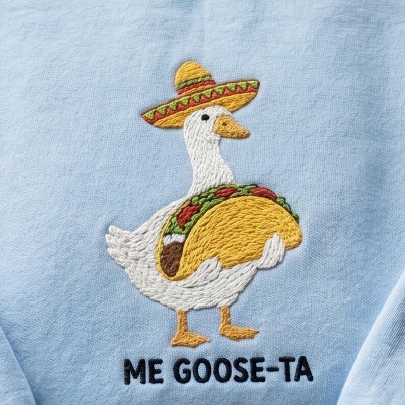 May include: Embroidered light blue sweatshirt featuring a white goose wearing a sombrero and a taco. The goose has a yellow beak and feet. The text "ME GOOSE-TA" is embroidered below the goose.