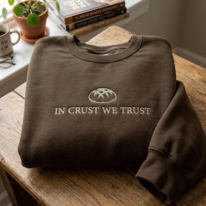 May include: A dark brown crewneck sweatshirt featuring an embroidered light green graphic of a round loaf of bread centered above the text IN CRUST WE TRUST in matching light green thread. The garment has long sleeves and a ribbed neckline.