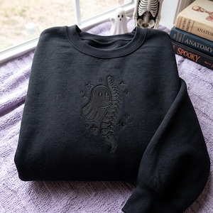 May include: A black crewneck sweatshirt featuring a tonal black embroidered design on the chest. The embroidery depicts a small ghost hugging a human spine, surrounded by several small four-pointed stars. The sweatshirt has long sleeves and ribbed cuffs.