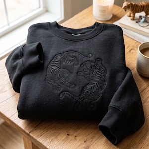 May include: A black long-sleeve sweatshirt featuring a tonal, embossed graphic of two tigers facing each other in a circular composition. The garment has a ribbed crew neckline and ribbed cuffs, showcasing detailed stitching of the tiger figures.