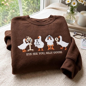 Könnte beinhalten: Ein dunkelbraunes Sweatshirt mit Rundhalsausschnitt und einer Stickerei von vier weißen Gänsen mit orangefarbenen Schnäbeln und Füßen. Die Gänse sind mit Sehtest-Ausrüstung dargestellt, darunter der Text "EYE SEE YOU, SILLY GOOSE" in weißer Schrift.