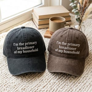 May include: Two baseball caps, one black and one brown, both featuring the white embroidered text "I'm the primary breadloser of my household" on the front panel. Each cap has a structured crown, a curved brim, and a washed, vintage-style fabric finish.