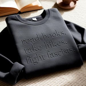 Puede incluir: Sudadera gris oscuro con la frase en relieve: "read books. take hikes. fight fascists." La prenda doblada descansa sobre una superficie texturizada, con un libro y una taza en el fondo.
