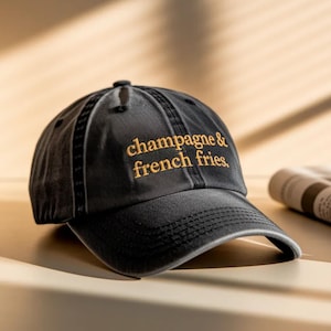 May include: A black baseball cap with the embroidered text "champagne & french fries" in gold thread. The cap has a curved brim and a distressed, vintage look.