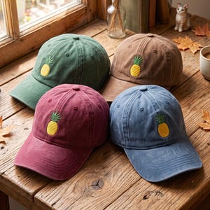 Op de afbeelding: Vier baseballpetten in groen, bruin, bordeauxrood en blauw, elk voorzien van een geborduurde gele ananas met groene bladeren op het voorpaneel. De petten hebben een gestructureerde kroon, een gebogen klep en een knoop aan de bovenkant.