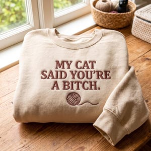 Pode incluir: Uma camisola de gola redonda bege com texto bordado em castanho escuro que diz MY CAT SAID YOU'RE A BITCH. Abaixo do texto encontra-se um novelo de lã castanho escuro bordado com um fio solto, centrado na parte frontal da peça.