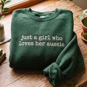 Könnte beinhalten: Ein dunkelwaldgrüner Rundhals-Pullover mit dem gestickten weißen Text "just a girl who loves her aussie" mittig auf der Brust. Das Kleidungsstück hat lange Ärmel, einen gerippten Halsausschnitt und eine weiche, innen angeraute Textur.