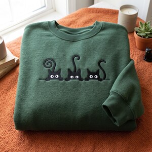 Puede incluir: Una sudadera de cuello redondo color verde bosque con un diseño bordado de tres gatos negros asomándose sobre una línea horizontal. Cada gato tiene ojos blancos grandes y una cola de forma distinta, una enroscada, una recta y una curva hacia un lado.
