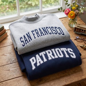 May include: A light gray crewneck sweatshirt with the text SAN FRANCISCO in dark blue lettering stacked on top of a navy blue sweatshirt with the text PATRIOTS in white lettering. Both garments feature a classic athletic font style.