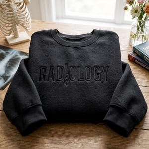 May include: A black crewneck sweatshirt featuring the word RADIOLOGY embroidered in a matching black thread across the chest. The letter I is replaced by a vertical graphic of a human spine, creating a cohesive design on the dark fabric.