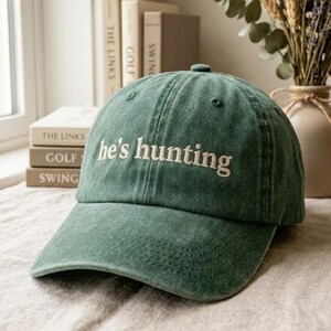 May include: A forest green baseball cap with a structured crown and a curved brim. The front of the cap features the embroidered text "he's hunting" in a light cream-colored font. The fabric has a slightly faded, washed-out appearance.