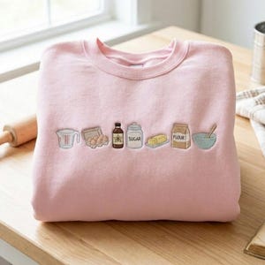 Op de afbeelding: Een lichtroze sweater met ronde hals voorzien van een geborduurde rij bakingrediënten, waaronder een maatbeker, een doos eieren, een flesje vanille-extract, een suikerpot, een blokje boter, een zak meel en een mengkom.